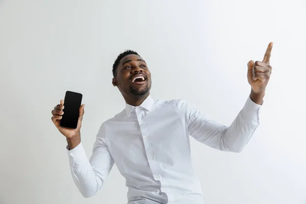 Innenporträt eines attraktiven jungen schwarzafrikanischen Mannes isoliert auf rosa Hintergrund, leeres Smartphone in der Hand, lächelnd in die Kamera, Bildschirm zeigend, glücklich und überrascht. menschliche Emotionen, Gesichtsausdruck — Stockfoto