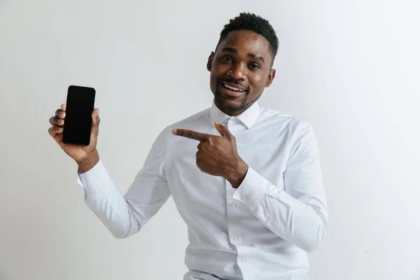 Retrato interior de un atractivo joven africano negro aislado sobre fondo rosa, sosteniendo un smartphone en blanco, sonriendo a la cámara, mostrando la pantalla, sintiéndose feliz y sorprendido. Emociones humanas, faciales — Foto de Stock