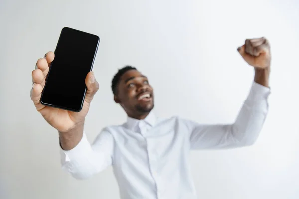 Innenporträt eines attraktiven jungen schwarzafrikanischen Mannes isoliert auf rosa Hintergrund, leeres Smartphone in der Hand, lächelnd in die Kamera, Bildschirm zeigend, glücklich und überrascht. menschliche Emotionen, Gesichtsausdruck — Stockfoto