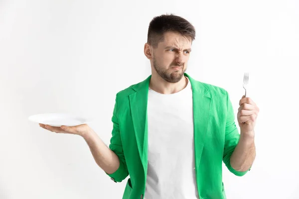 Young sad attractive guy holding empty dish and fork isolated on grey background. — Stock Photo, Image