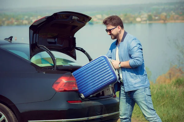 英俊的微笑男子去度假，装他的手提箱在汽车后备箱 — 图库照片