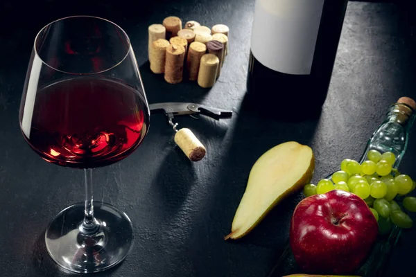 Una bottiglia e un bicchiere di vino rosso con frutta su sfondo pietra scura — Foto Stock