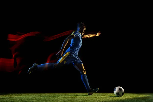 Male soccer player kicking ball on dark background in mixed light