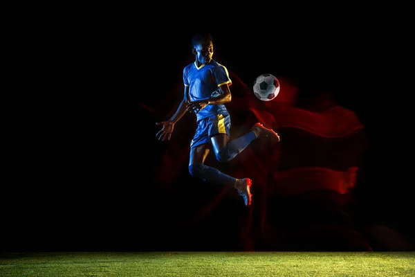 Male soccer player kicking ball on dark background in mixed light
