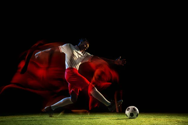 Male soccer player kicking ball on dark background in mixed light