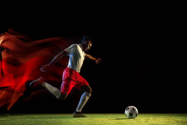 Male soccer player kicking ball on dark background in mixed light