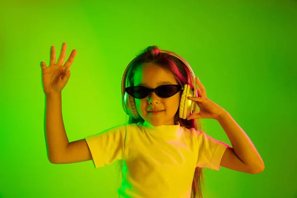 Portret van klein meisje in koptelefoon op groene achtergrond in neon licht — Stockfoto