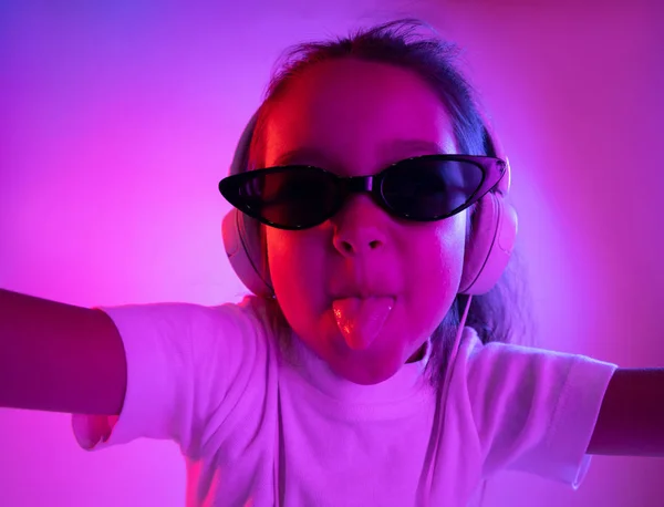 Portret małej dziewczynki w słuchawkach na fioletowym tle gradientu w świetle neonowym — Zdjęcie stockowe