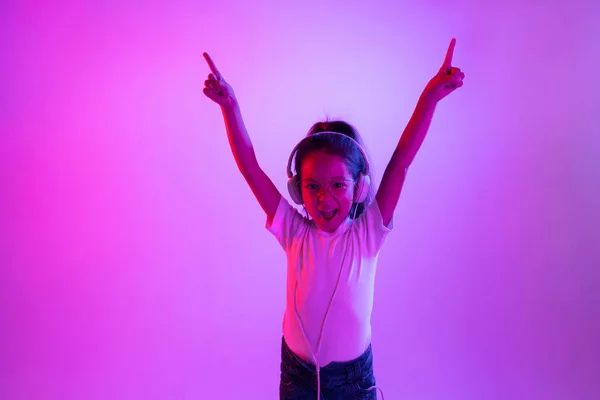 Portret małej dziewczynki w słuchawkach na fioletowym tle gradientu w świetle neonowym — Zdjęcie stockowe