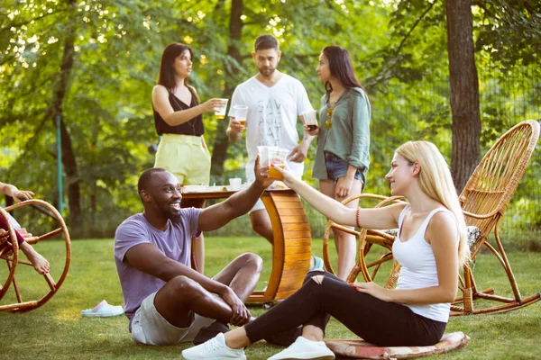 Счастливые друзья едят и пьют пиво за ужином с барбекю на закате — стоковое фото