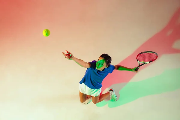 Young woman in blue shirt playing tennis in mixed light. Youth, flexibility, power and energy.