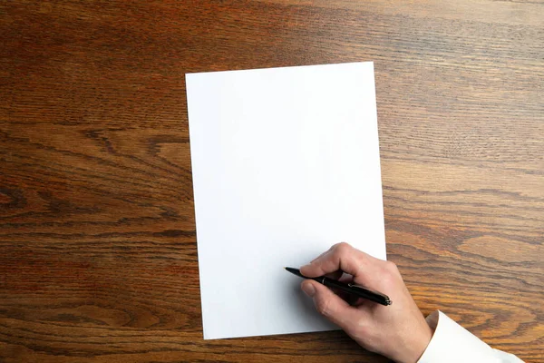 Mão masculina segurando caneta e escrevendo em folha vazia em fundo de madeira para texto ou design — Fotografia de Stock