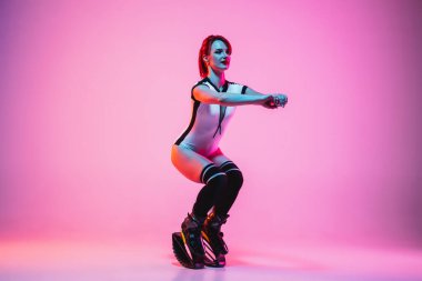 Beyaz spor giysili güzel kızıl saçlı bir kadın kangoya atlıyor ve neon ışıklarıyla izole edilmiş bir şekilde stüdyoda ayakkabılarını giyiyor.