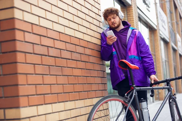 Bonito jovem usando telefone celular e fones de ouvido enquanto estava perto de sua bicicleta ao lado dele — Fotografia de Stock