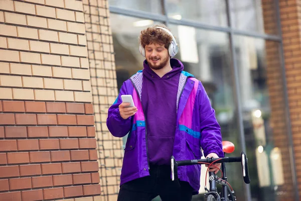 隣の自転車の近くに立っている間に携帯電話とヘッドフォンを使っているハンサムな若い男 — ストック写真