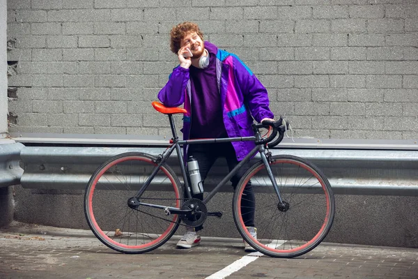 Bonito jovem usando telefone celular e fones de ouvido enquanto estava perto de sua bicicleta ao lado dele — Fotografia de Stock