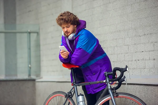 Bonito jovem usando telefone celular e fones de ouvido enquanto estava perto de sua bicicleta ao lado dele — Fotografia de Stock