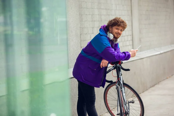 Bonito jovem usando telefone celular e fones de ouvido enquanto estava perto de sua bicicleta ao lado dele — Fotografia de Stock