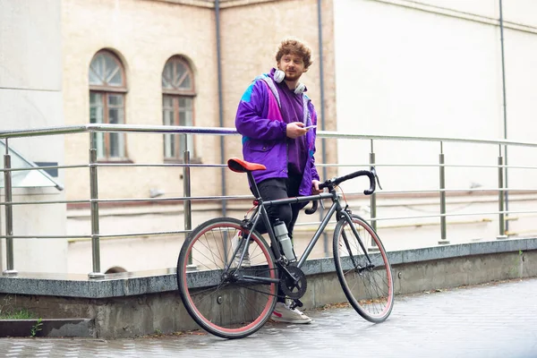 Beau jeune homme utilisant téléphone portable et écouteurs tout en se tenant près de son vélo à côté de lui — Photo