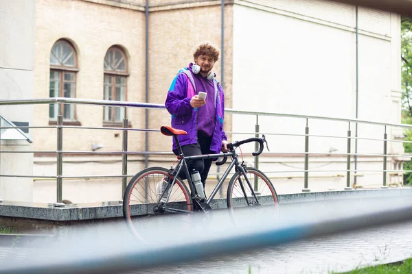 Hezký mladý muž pomocí mobilního telefonu a sluchátek, zatímco stojí u svého kola vedle něj — Stock fotografie