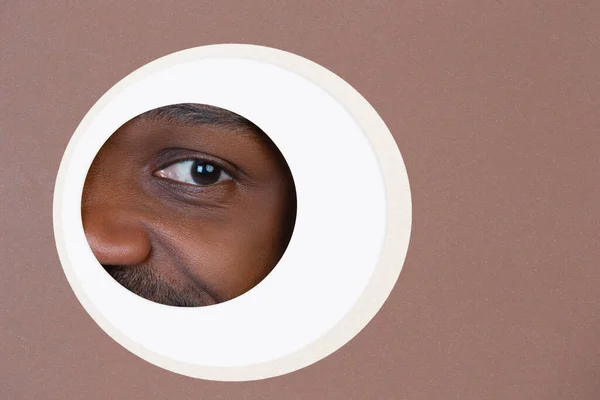 Eye of smiling african-american man peeking throught circle in brown background — Stock Photo, Image