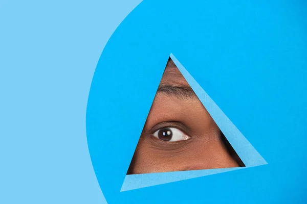 Eye of african-american man peeking throught triangle in blue background