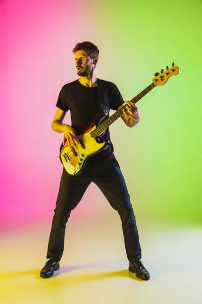 Jovem músico caucasiano tocando baixo em luz de néon sobre fundo rosa-verde — Fotografia de Stock