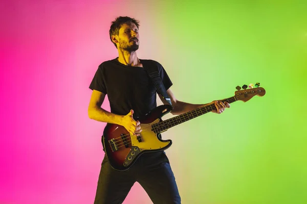 Young caucasian musician playing bass guitar in neon light on pink-green background — Stock Photo, Image