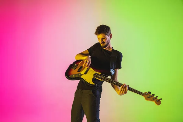 Jovem músico caucasiano tocando baixo em luz de néon sobre fundo rosa-verde — Fotografia de Stock
