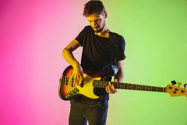 Junger kaukasischer Musiker spielt Bassgitarre im Neonlicht auf rosa-grünem Hintergrund — Stockfoto