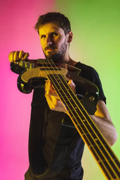 Young caucasian musician playing bass guitar in neon light on pink-green background — Stock Photo, Image