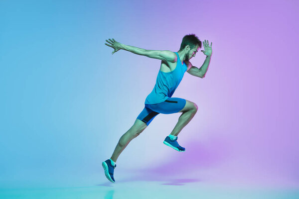 Full length portrait of active young caucasian running, jogging man on gradient studio background in neon light