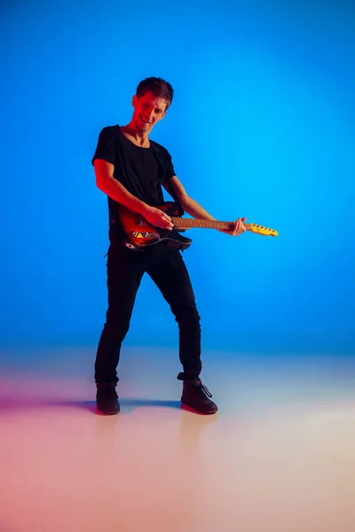 Jovem músico caucasiano tocando guitarra em luz de néon em fundo azul, inspirado — Fotografia de Stock