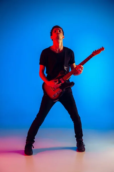 Young caucasian musician playing guitar in neon light on blue background, inspired