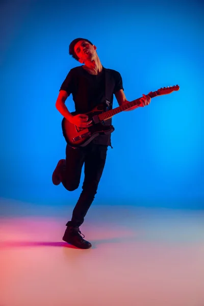 Jovem músico caucasiano tocando guitarra em luz de néon em fundo azul, inspirado — Fotografia de Stock