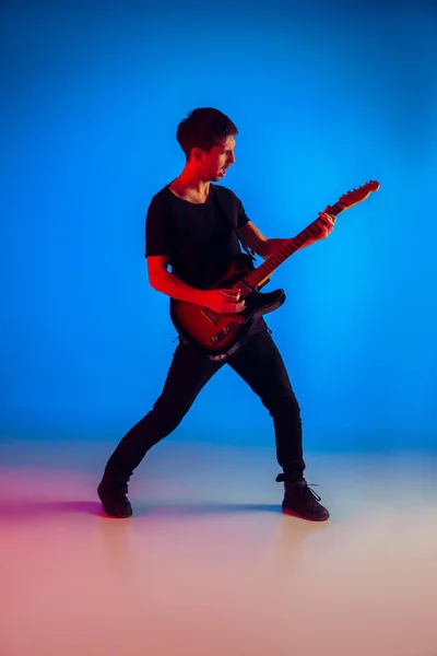 Joven músico caucásico tocando la guitarra en luz de neón sobre fondo azul, inspirado — Foto de Stock