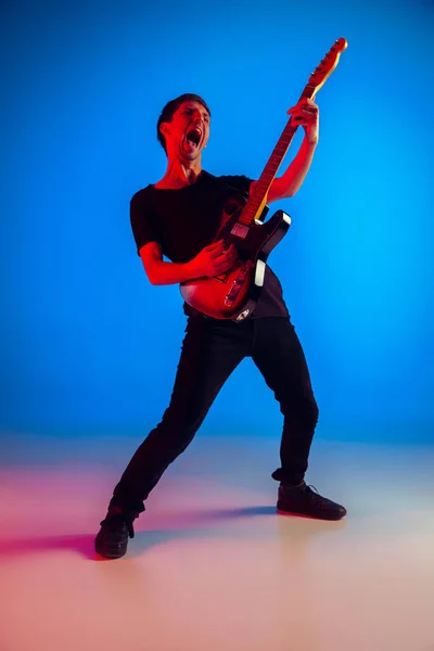 Jovem músico caucasiano tocando guitarra em luz de néon em fundo azul, inspirado — Fotografia de Stock