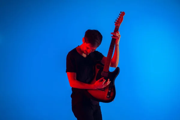 Молодий кавказький музикант грає на гітарі в неоновому світлі на синьому фоні, натхненний — стокове фото