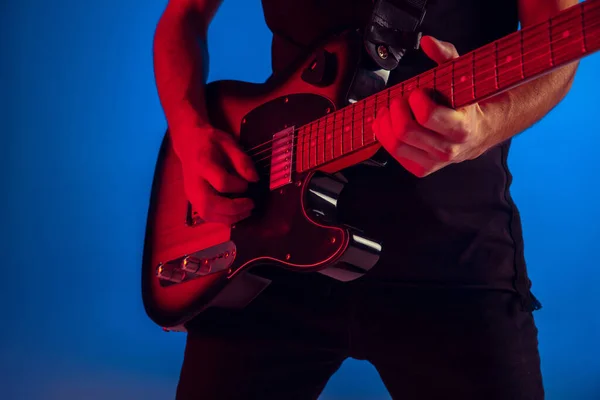 Young caucasian musician playing guitar in neon light on blue background, inspired