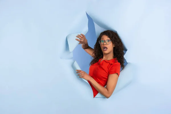 Vrolijke jonge vrouw poseert in gescheurd blauw papier gat achtergrond, emotioneel en expressief — Stockfoto