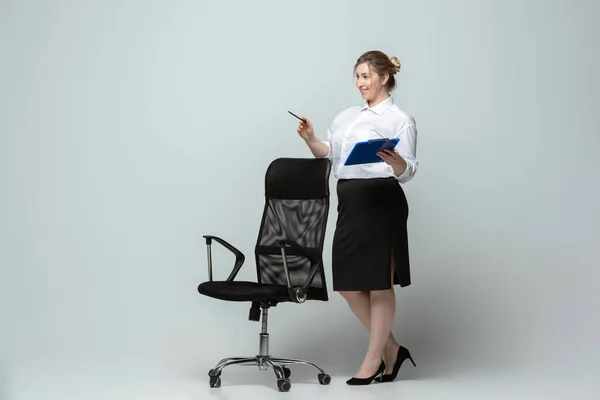Mladá běloška v kanceláři se obléká na šedé pozadí. Tělesně pozitivní ženský charakter. plus velikost podnikatelka — Stock fotografie