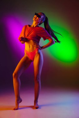 Güzel, baştan çıkarıcı bir kız, modaya uygun mayo giymiş, neon ışıklı, iki renkli stüdyo arka planında. Yaz, tatil köyü, moda ve hafta sonu konsepti