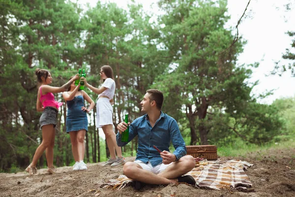 Group of friends clinking beer bottles during picnic in summer forest. Lifestyle, friendship
