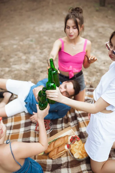 Група друзів смердить пляшками пива під час пікніка в літньому лісі. Стиль життя, дружба — стокове фото