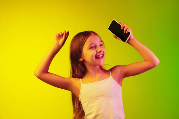 Kleine kaukasische Mädchen porträtieren isoliert auf einem Gradienten-Hintergrund im Neonlicht. Konzept menschlicher Emotionen, Mimik, moderner Geräte und Technologien. Kopierraum. — Stockfoto