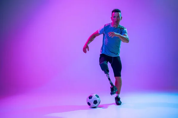 Athlete with disabilities or amputee isolated on gradient studio background. Professional male football player with leg prosthesis training and practicing in studio. — Stock Photo, Image