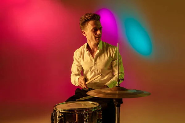 Junger kaukasischer Musiker, Schlagzeuger, der auf einem Gradienten-Hintergrund im Neonlicht spielt. Konzept Musik, Hobby, Festival — Stockfoto