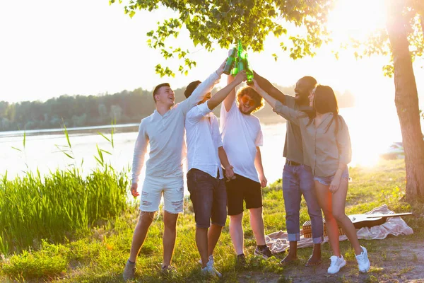 Group of friends clinking beer bottles during picnic at the beach. Lifestyle, friendship, having fun, weekend and resting concept.