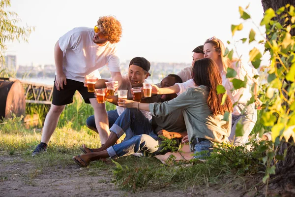 Grupo de amigos tintineando vasos de cerveza durante el picnic en la playa. Estilo de vida, amistad, diversión, fin de semana y concepto de descanso. — Foto de Stock