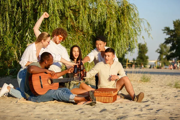 Grupo de amigos tintineando vasos de cerveza durante el picnic en la playa. Estilo de vida, amistad, diversión, fin de semana y concepto de descanso. — Foto de Stock